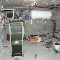Kotłownia w piwnicy z kotłem na paliwo stałe marki ECO, bojlerem i instalacją hydrauliczną z rur miedzianych i plastikowych.
