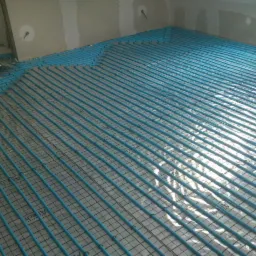 Niebieskie rurki systemu rozprowadzenia ciepła ułożone równolegle na siatce podłogowej w surowym stanie, przygotowane do zalania wylewką.