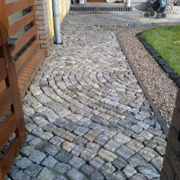 Ścieżka z kostki granitowej ułożona w łukowaty wzór, prowadząca od drewnianej bramy do wejścia domu. Obok ścieżki widoczny pas żwiru i trawnik.