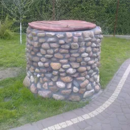 Kamienna studnia kręgowa z drewnianym wiekiem na trawniku, obok ścieżki z kostki brukowej, otoczona zielenią.