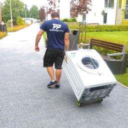 MR TRANS Małgorzata Rypień (Grupa MR Trans Przeprowadzki Warszawa) - Tragarz w niebieskiej koszulce z logo PP Przeprowadzki Polska transportuje białą pralkę na wózku z zielonymi kółkami po chodniku z kostki brukowej w otoczeniu zieleni i nowoczesnej zabudowy.