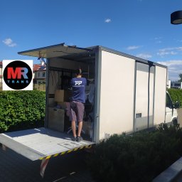 MR TRANS Małgorzata Rypień (Grupa MR Trans Przeprowadzki Warszawa) - Mężczyzna w koszulce z logo 'Przeprowadzki Polska' ładuje meble i kartony do meblowozu z otwartą windą załadowczą, podczas przeprowadzki w słoneczny dzień.