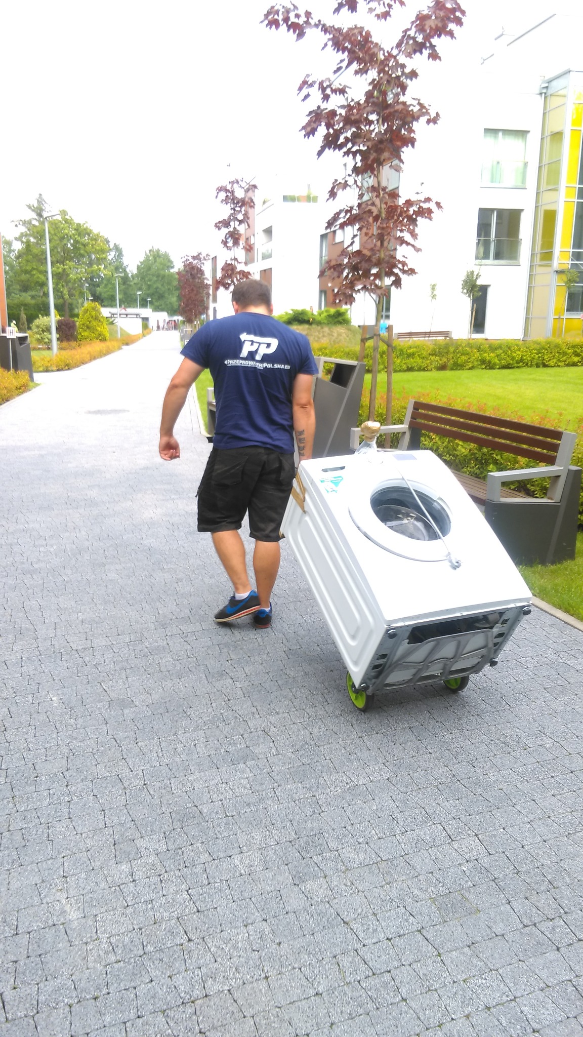 Tragarz w niebieskiej koszulce z logo PP Przeprowadzki Polska transportuje białą pralkę na wózku z zielonymi kółkami po chodniku z kostki brukowej w otoczeniu zieleni i nowoczesnej zabudowy.