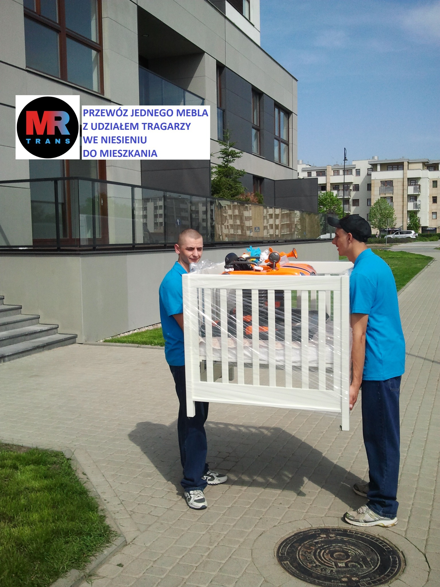 Dwóch mężczyzn w niebieskich koszulkach niesie zapakowane w folię białe łóżeczko dziecięce na tle nowoczesnego budynku mieszkalnego.