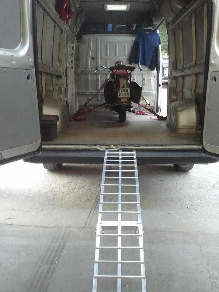 Wnętrze busa z otwartymi drzwiami, widoczny skuter zabezpieczony pasami transportowymi i rampa najazdowa, przygotowany do transportu.