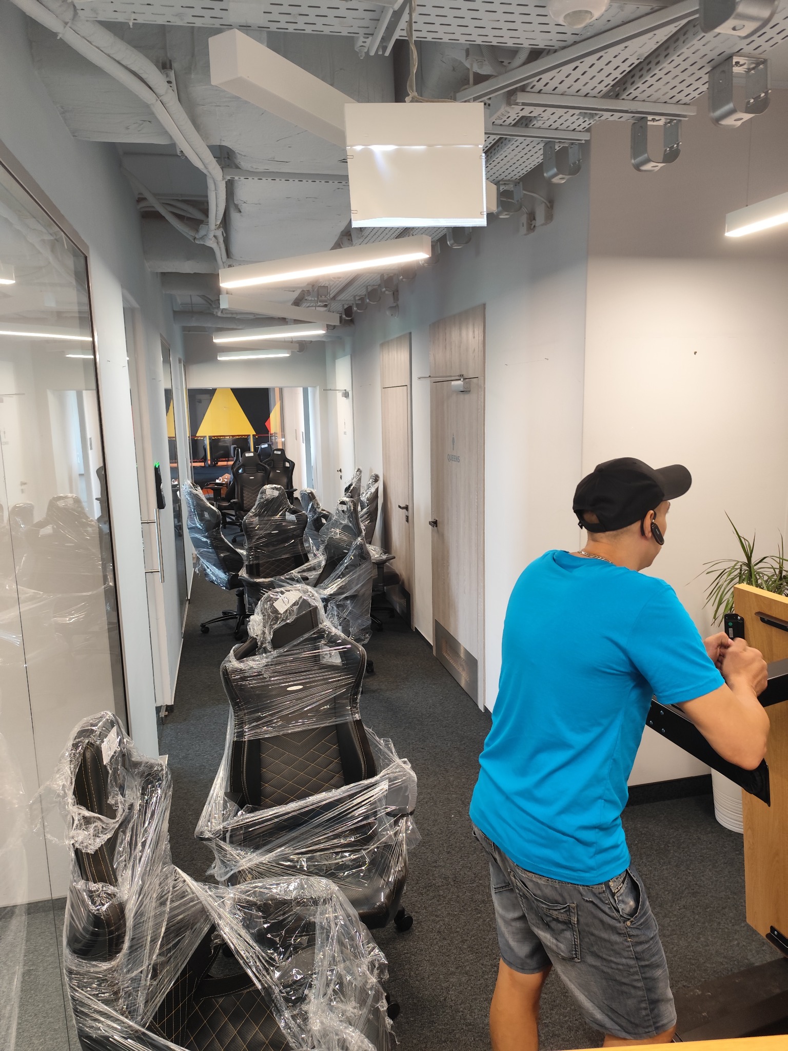Korytarz biurowy z krzesłami biurowymi w folii, przygotowanymi do transportu, widoczny mężczyzna w niebieskiej koszulce i czapce, prawdopodobnie pracownik firmy przeprowadzkowej.