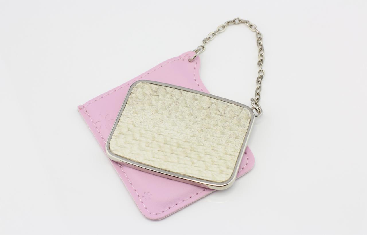 Kompaktowe lusterko z łańcuszkiem, leżące na różowym, skórzanym etui z wytłoczonym kwiatkiem, na białym tle.