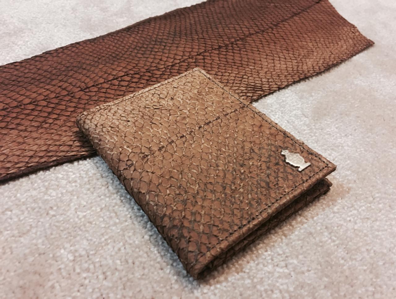 Portfel z brązowej skóry o fakturze łusek z metalowym logo w kształcie butelki na jasnym dywanie, obok fragment skóry w tym samym stylu.