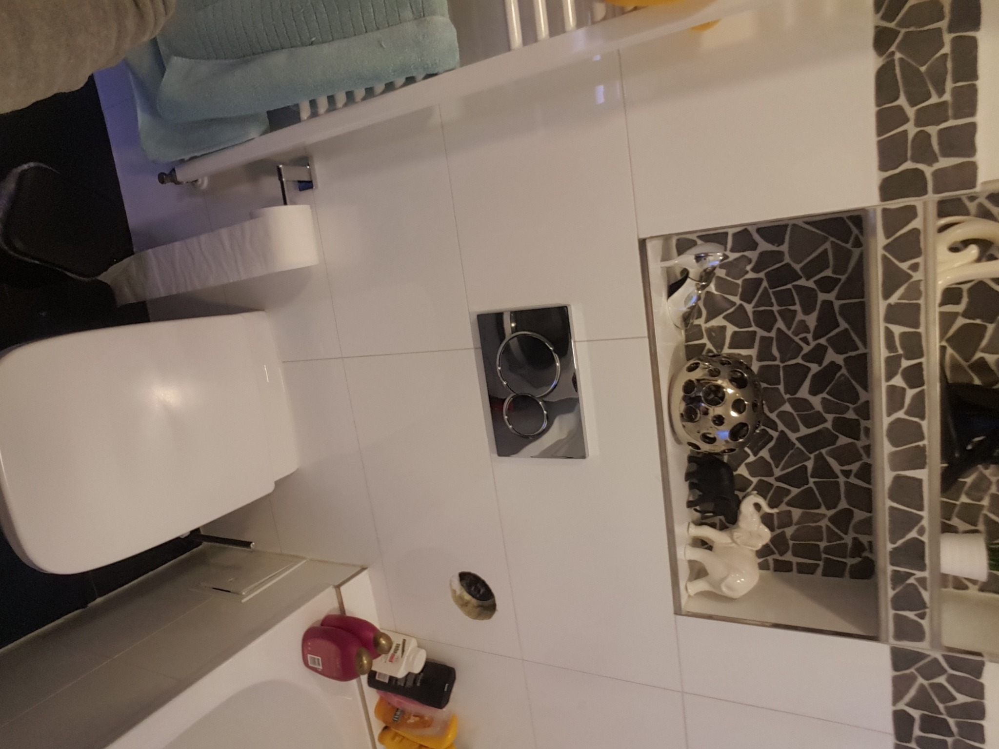 Nowoczesna łazienka z białą ceramiką, mozaiką w odcieniach szarości, wiszącą miską WC, chromowanym przyciskiem spłukującym i dekoracyjną niszą z figurkami.