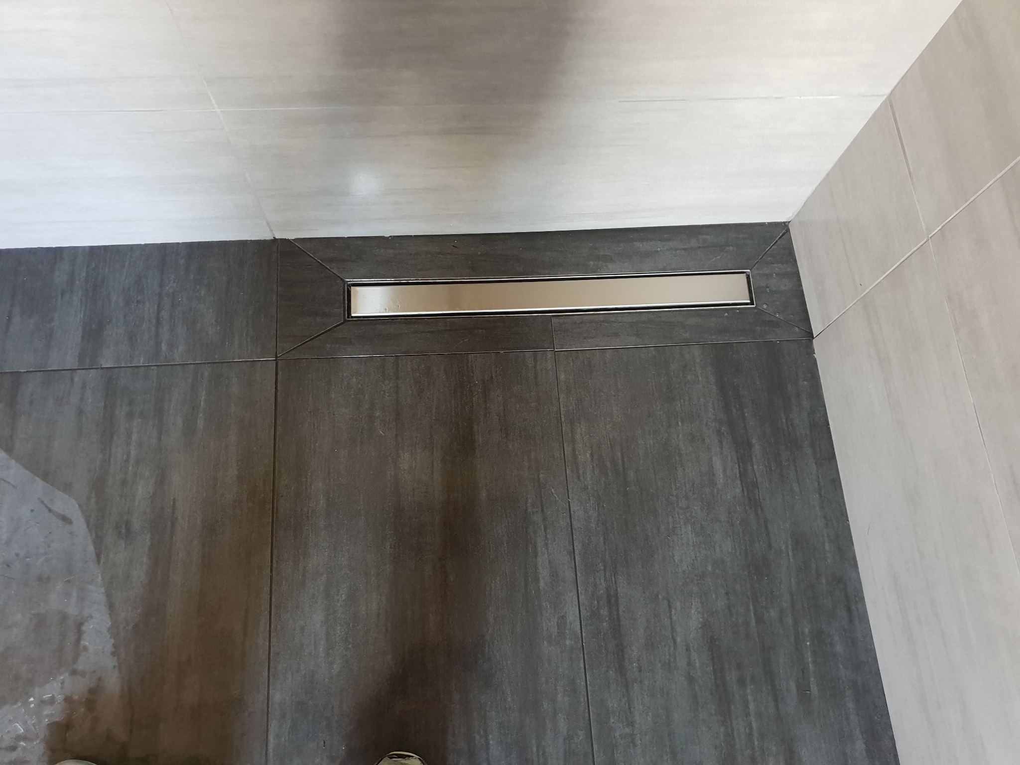 Nowoczesna łazienka z ciemnoszarą podłogą wyłożoną płytkami imitującymi drewno i odpływem liniowym ze stali nierdzewnej, widoczne fragmenty jasnych płytek ściennych.