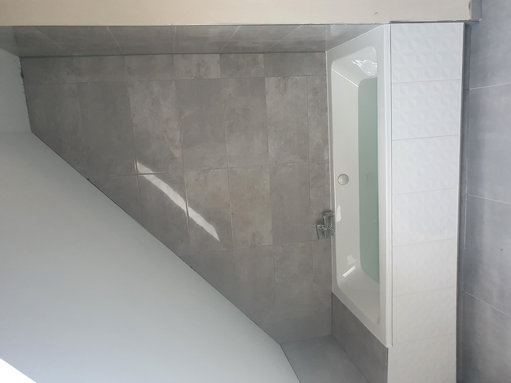 Nowoczesna, biała wanna z chromowaną baterią, otoczona szarymi płytkami ceramicznymi o różnych odcieniach i fakturach, w nowo wykończonej łazience.