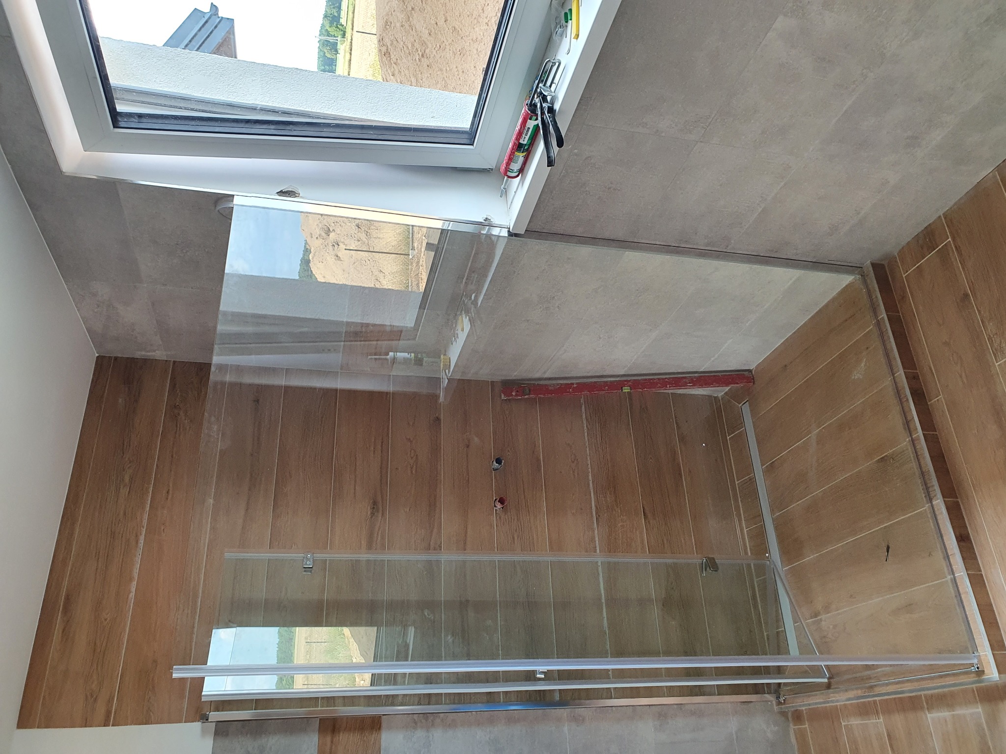 Szklana kabina prysznicowa montowana w łazience z oknem dachowym, widoczne narzędzia montażowe i poziomica.