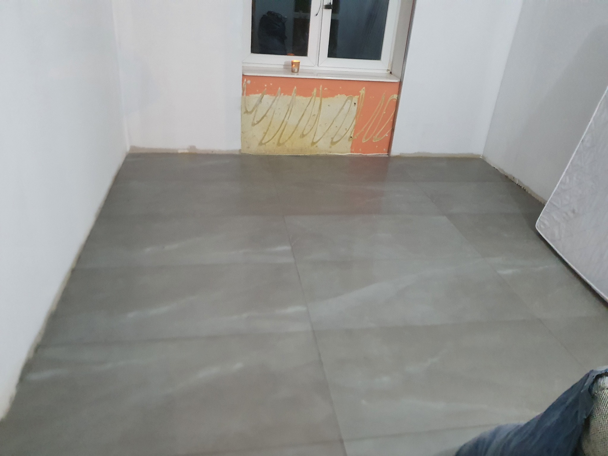 Świeżo ułożone, szare płytki podłogowe w pomieszczeniu w trakcie remontu, widoczne ślady po kleju pod oknem i fragment materaca oparty o ścianę.