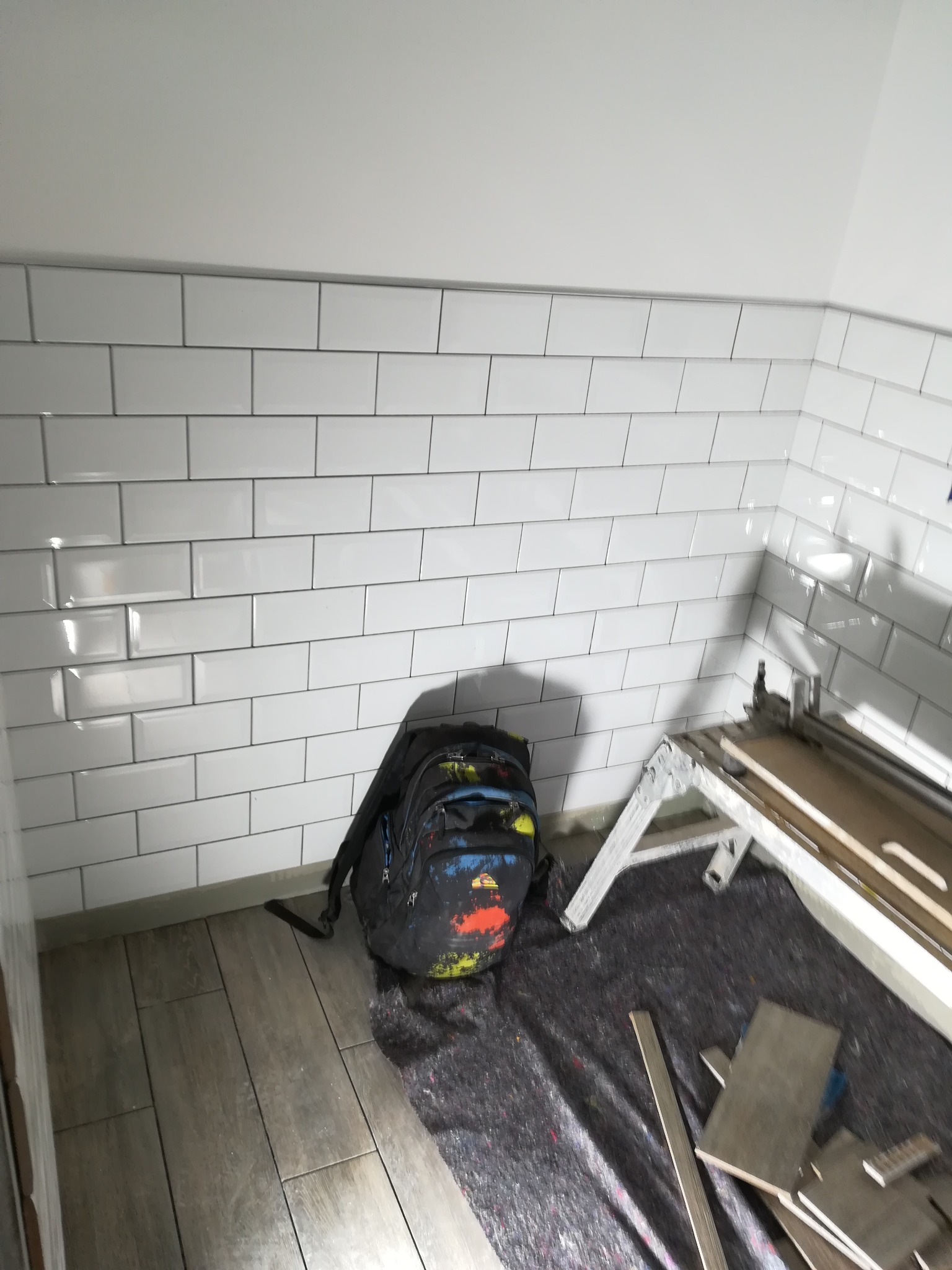 Świeżo położone, błyszczące, białe płytki ścienne typu metro w łazience, z widocznym plecakiem i gilotyną do cięcia płytek na podłodze imitującej drewno.
