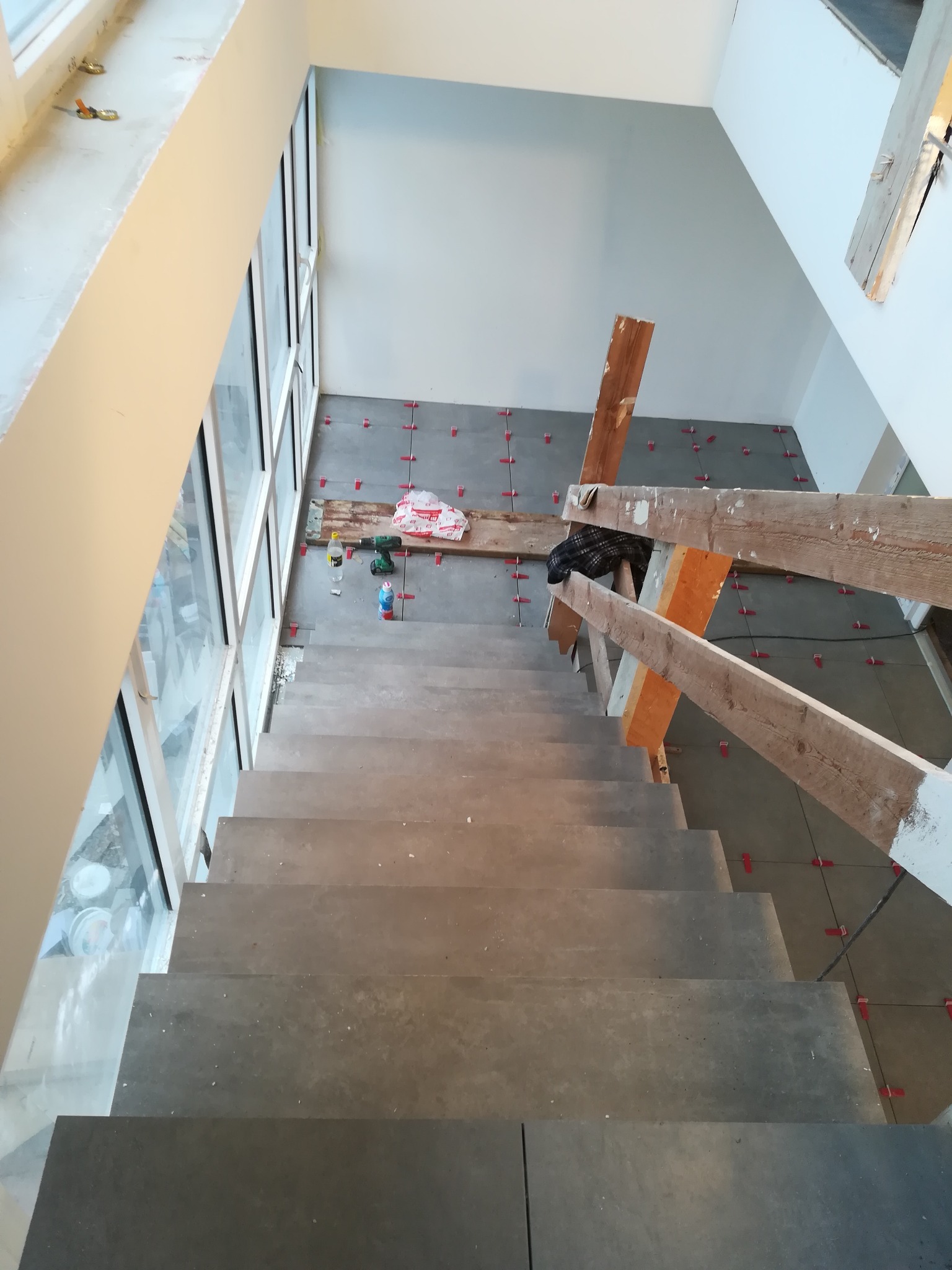 Montaż szarych płytek na schodach wewnątrz domu, widoczne narzędzia i poziomica, na ścianie widoczne czerwone krzyżyki dystansowe.