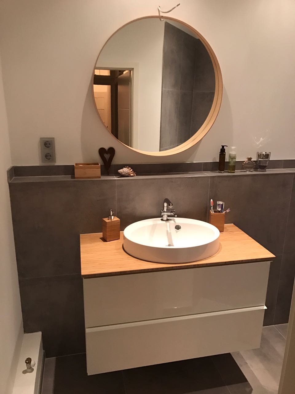 Nowoczesna łazienka z okrągłym lustrem w drewnianej ramie, białą umywalką nablatową na szafce z dwoma szufladami i drewnianym blatem, szara glazura na ścianie.