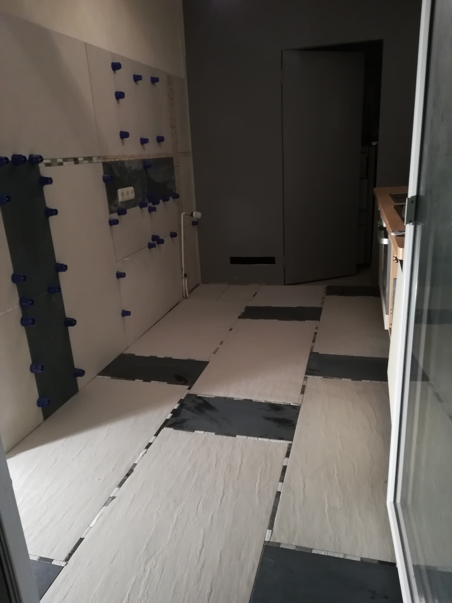 Układanie płytek w kuchni: białe płytki na ścianach z czarnymi akcentami, niebieskie krzyżyki dystansowe, częściowo ułożona podłoga z szarych i czarnych płytek.