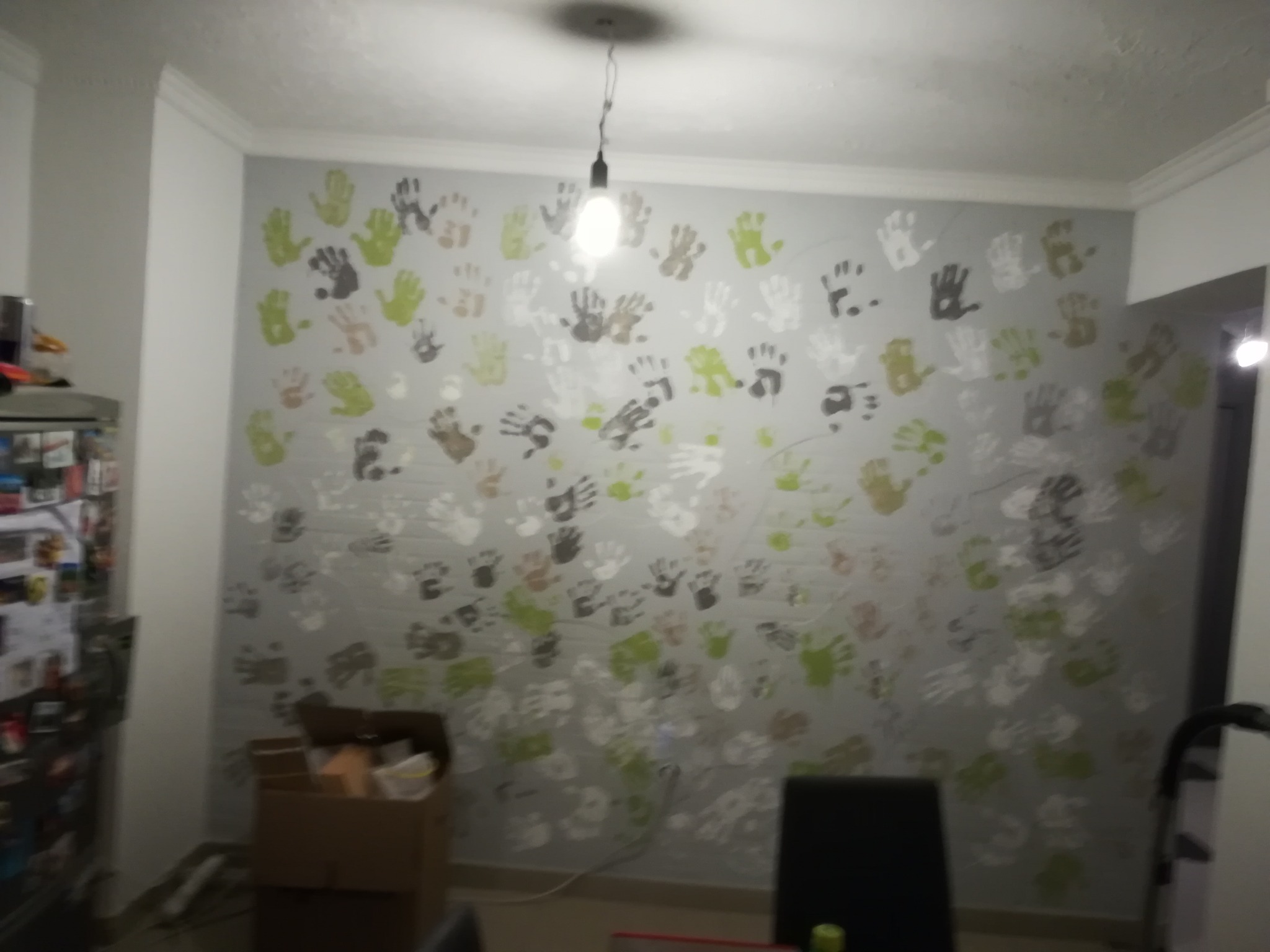 Ściana w pokoju pomalowana w odciski dłoni w różnych kolorach: zielonym, brązowym, szarym i białym, podświetlona pojedynczą żarówką.