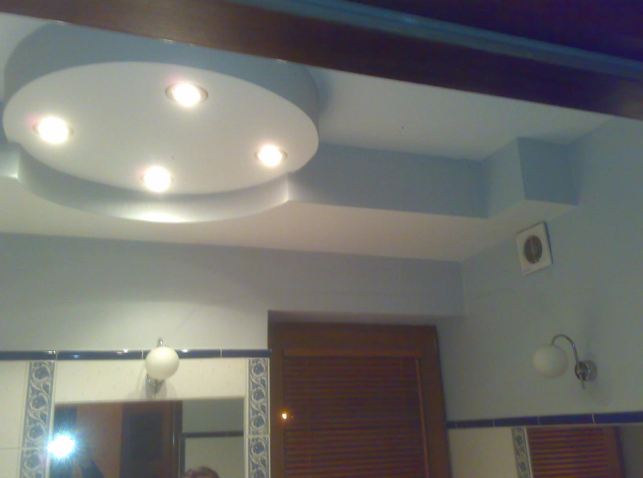 Okrągły podwieszany sufit z punktowym oświetleniem w łazience z niebieskimi ścianami, widoczne lustro z ozdobnymi płytkami i drewniane żaluzje.