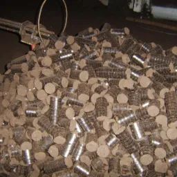 Stos brykietu z węgla brunatnego, cylindryczne kawałki z widocznymi śladami prasy, ułożone chaotycznie na ciemnym tle przemysłowym.