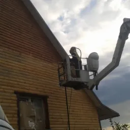 Pracownik w podnośniku koszowym czyści drewnianą elewację domu. Widoczne zabezpieczone okno i fragment białego samochodu dostawczego.