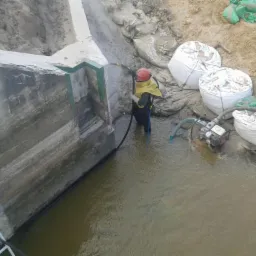 Pracownik w kombinezonie piaskuje betonową konstrukcję w wodzie, widoczne worki z piaskiem i pompa. Ujęcie z góry.