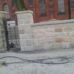 Kamienny mur z bramą i słupkami po renowacji, na tle ceglanego budynku. Na ziemi leżą węże i sprzęt. Widoczny szyld 'Obiekt Chroniony SOLID'.