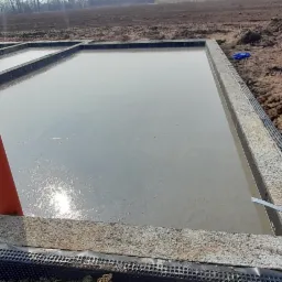 Zalany betonem fundament płyty w ziemi z wystającymi rurami wentylacyjnymi, widoczne ślady opon na ziemi.