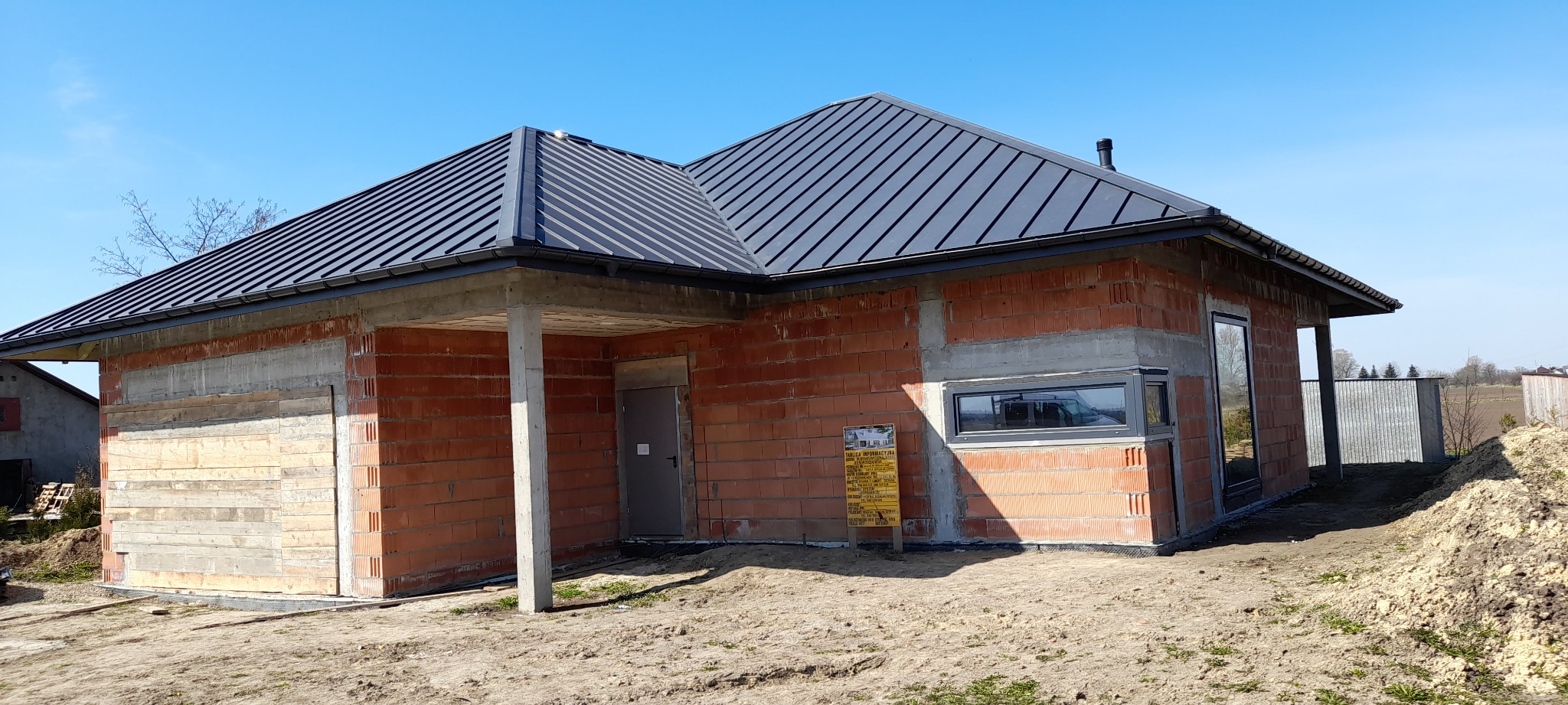 Dom murowany w trakcie budowy z czarnym dachem z blachy, widoczne ściany z czerwonej cegły, betonowe nadproża, okna, drzwi i tablica informacyjna na placu budowy.