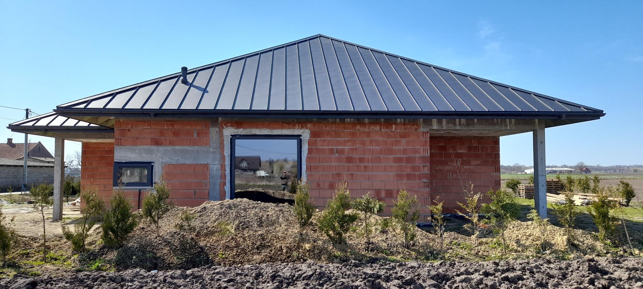 Surowy budynek z czerwonej cegły z zamontowaną ciemną blachodachówką i oknami, widoczne fundamenty i nasadzenia młodych drzewek iglastych.