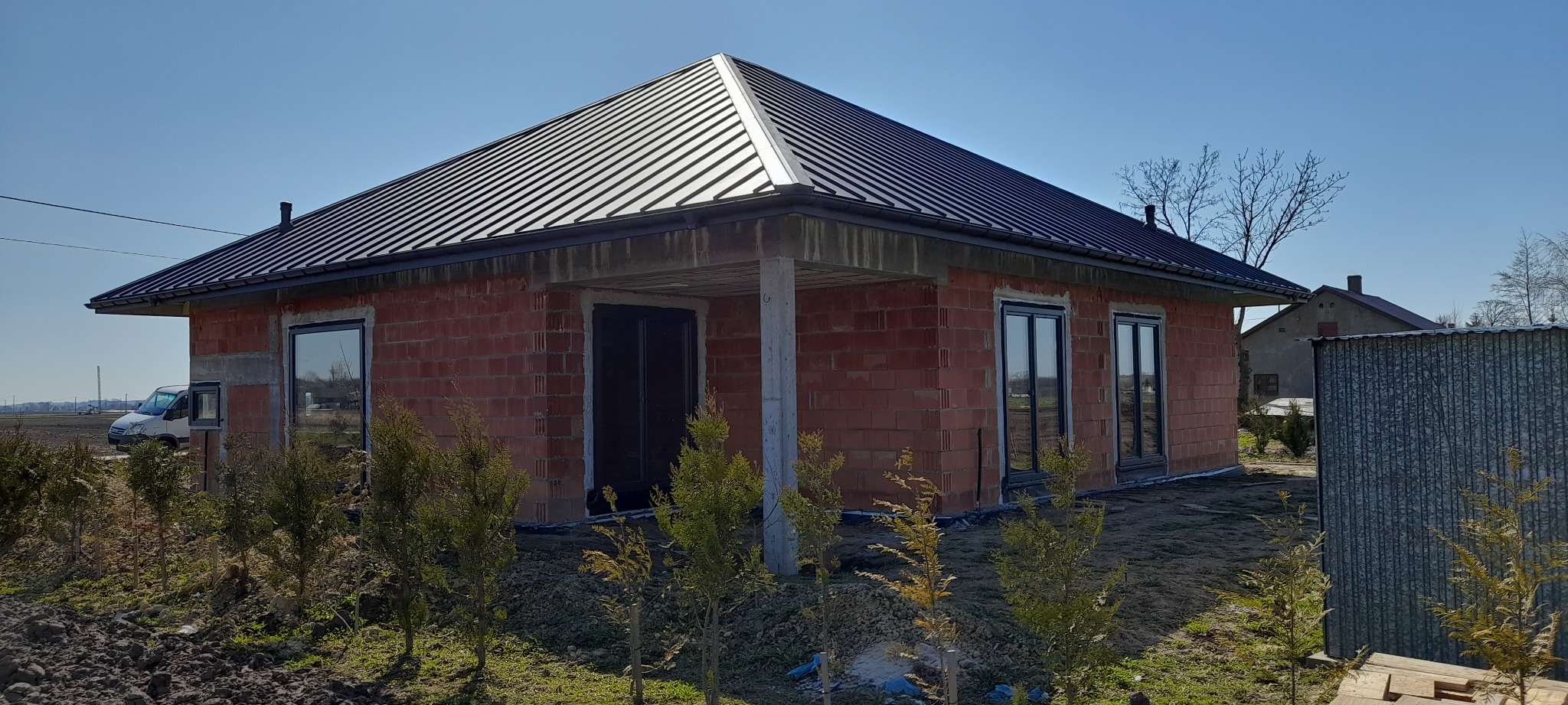 Surowy, parterowy dom z czerwonej cegły z czarnym, metalowym dachem, widoczne okna, betonowy filar i młode drzewka iglaste na pierwszym planie.