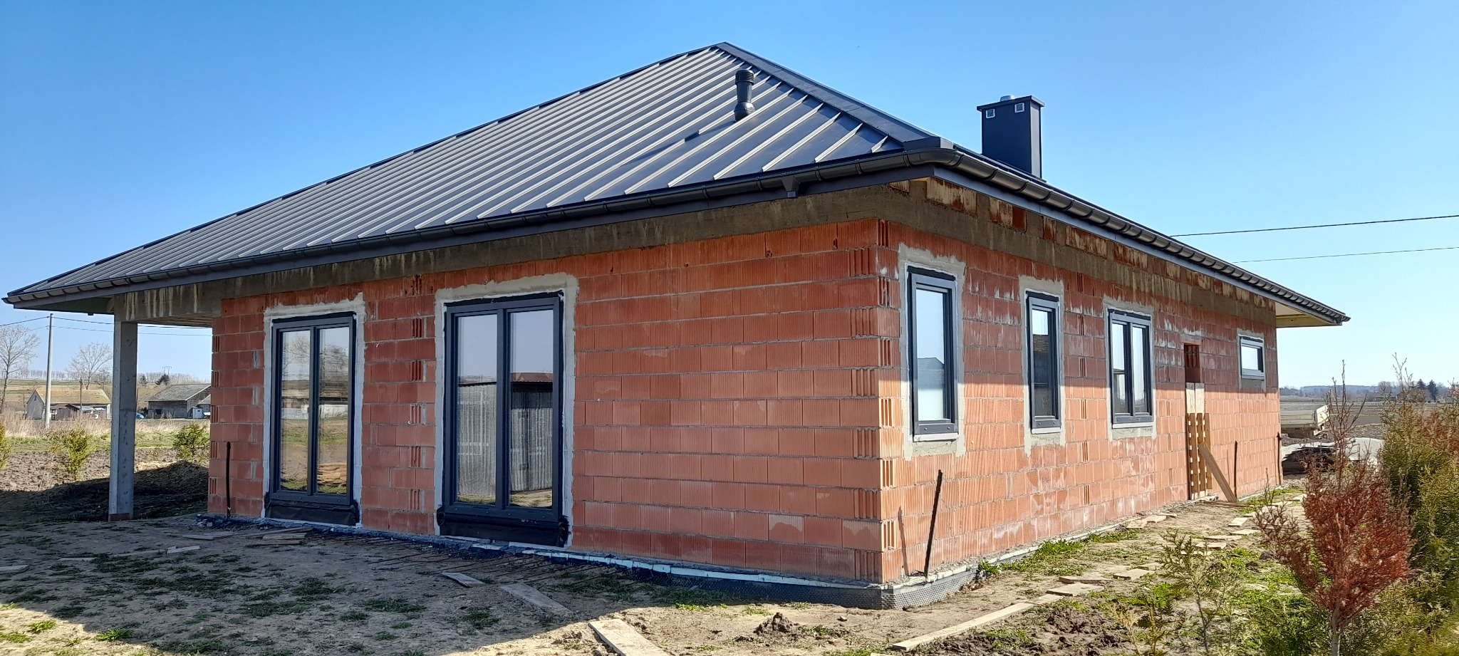 Surowy stan domu jednorodzinnego z czerwonej cegły z czarną blachodachówką, widoczne czarne ramy okienne, brak elewacji, teren wokół domu częściowo uporządkowany.