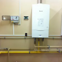 Dwa białe, wiszące piece gazowe marki Junkers, z widocznymi rurami gazowymi i wodnymi oraz elementami instalacji elektrycznej na beżowej ścianie.