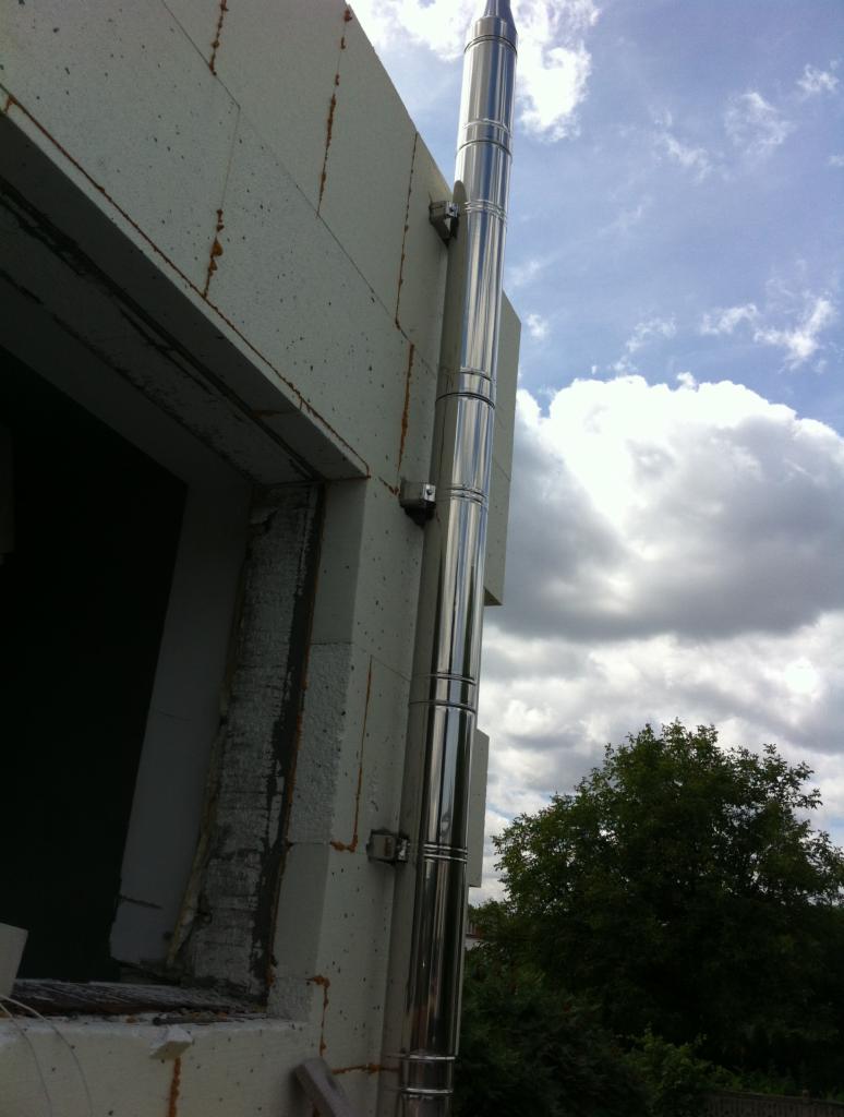 Zewnętrzny, metalowy komin spalinowy zamontowany przy ścianie budynku z betonu komórkowego, widoczne uchwyty mocujące i fragment okna.