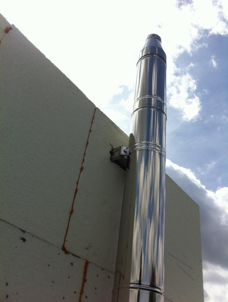 Błyszczący, metalowy komin przymocowany do ściany budynku z widocznym ociepleniem. Komin odbija błękit nieba i chmury. Ujęcie z dołu.