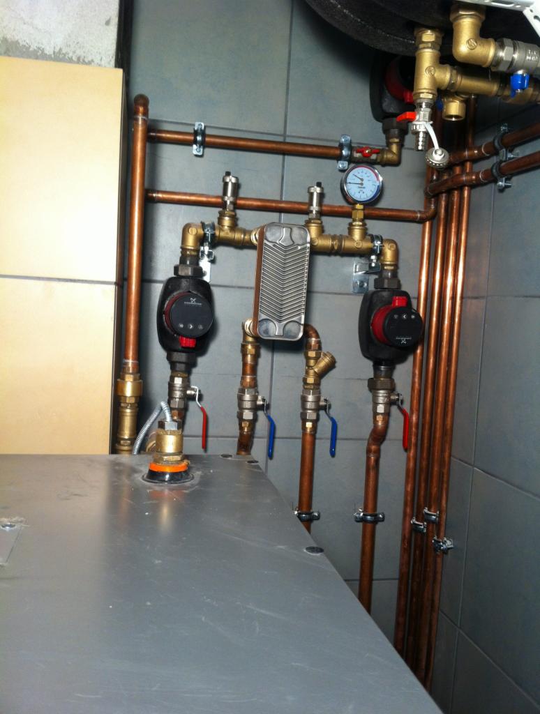 Miedziane rury instalacji grzewczej z zaworami, pompami obiegowymi i manometrem, zamontowane na szarej płytce w kotłowni. Widoczny fragment kotła.