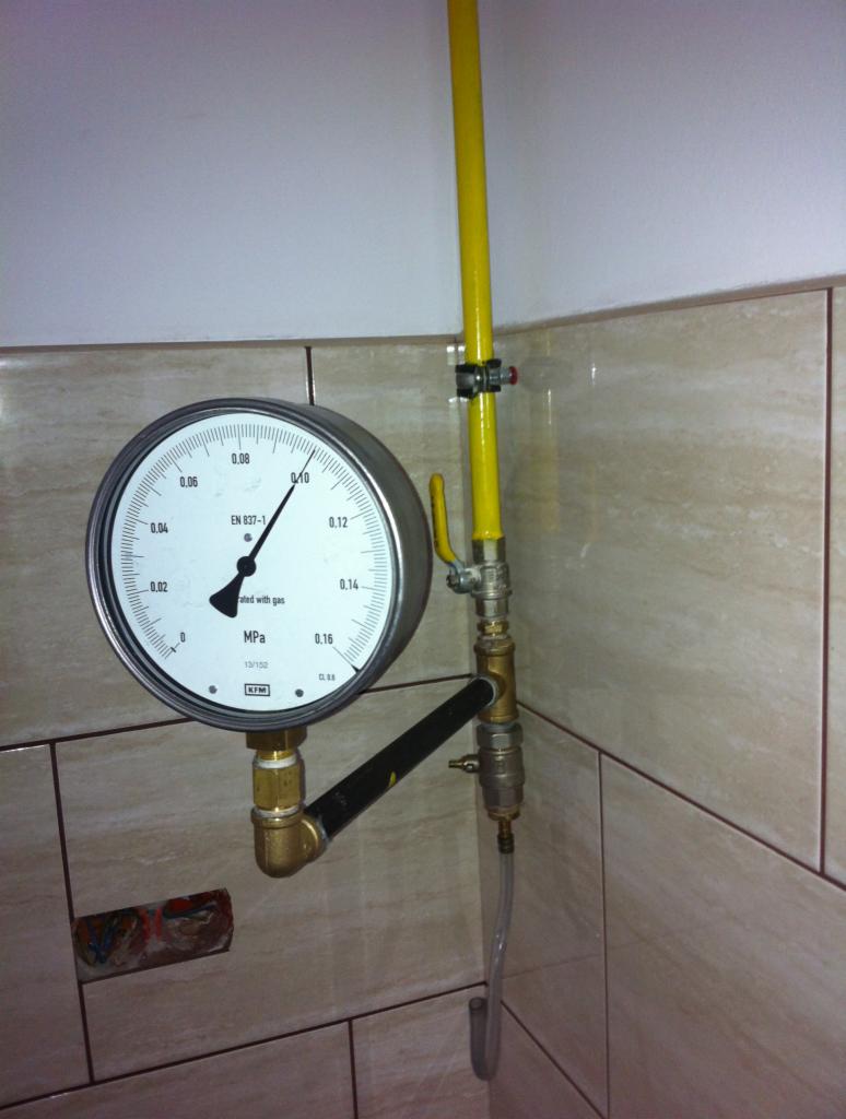 Manometr przy żółtej rurze gazowej na tle płytek. Instalacja gazowa z czujnikiem ciśnienia, widoczne złączki i zawór. Precyzyjny montaż instalacji gazowej w pomieszczeniu.