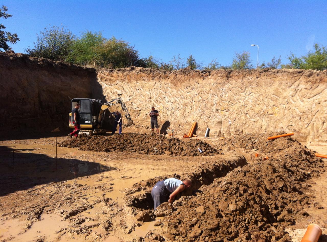Wykop pod instalacje: robotnicy pracują w wykopie, widoczny mały traktor, pomarańczowe rury. Teren budowy w słoneczny dzień.