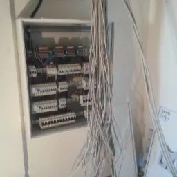 Otwarta skrzynka rozdzielcza z licznymi bezpiecznikami i wystającymi przewodami elektrycznymi, widoczne również fragmenty ścian i podłogi w trakcie remontu.