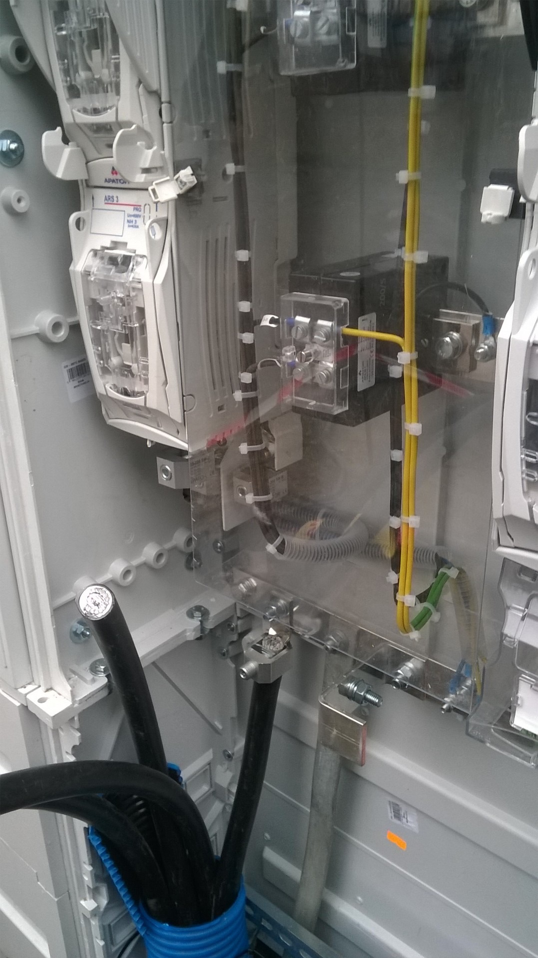 Wnętrze skrzynki elektrycznej z zamontowanymi bezpiecznikami, przewodami o różnych kolorach zabezpieczonymi opaskami i szynami uziemiającymi, widoczne elementy przezroczystej obudowy.