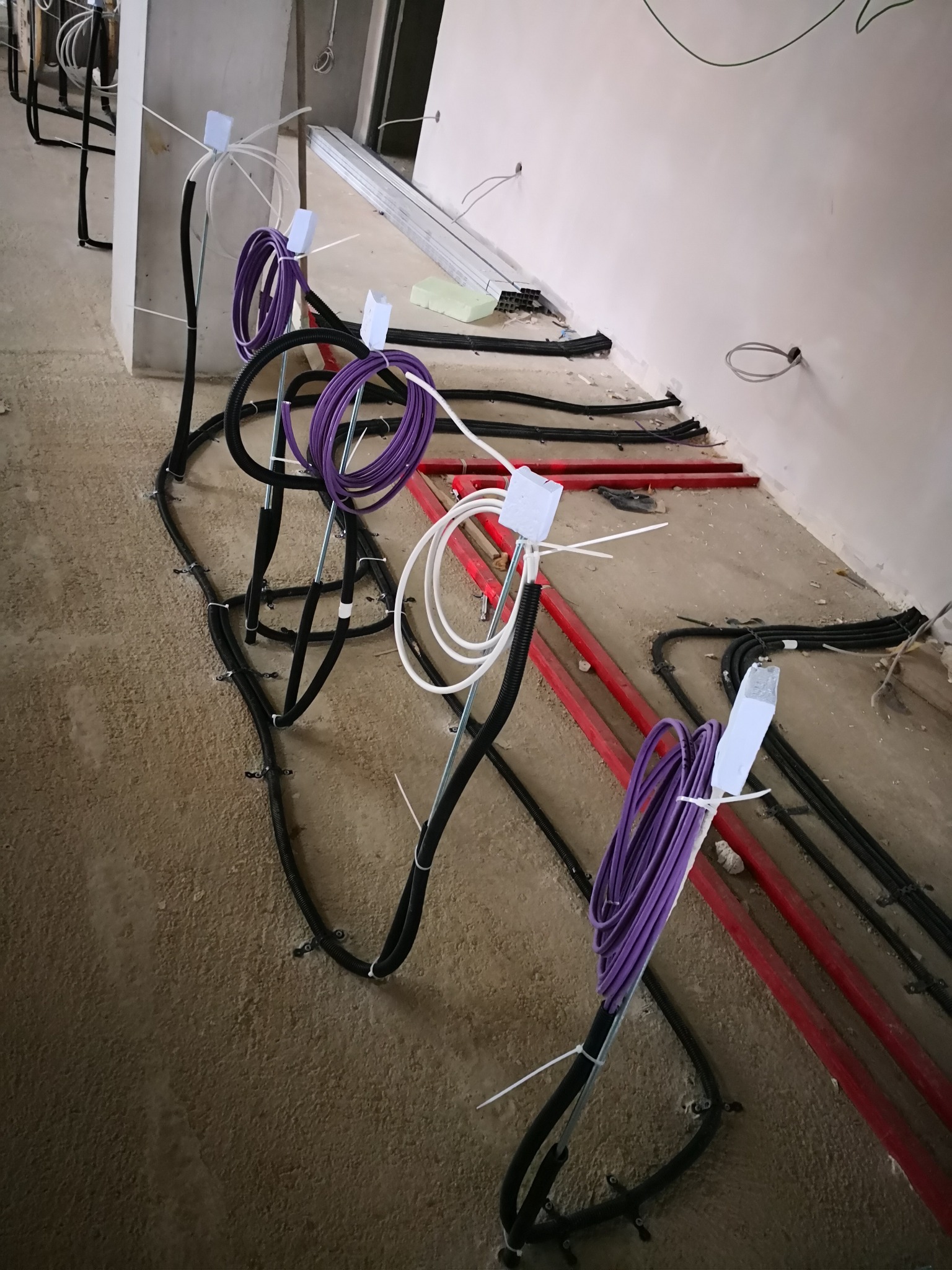 Kable elektryczne prowadzone po podłodze w trakcie instalacji; czarne rury karbowane mocowane do podłoża, obok czerwone rury grzewcze, zwinięte fioletowe i białe kable zabezpieczone opaskami...