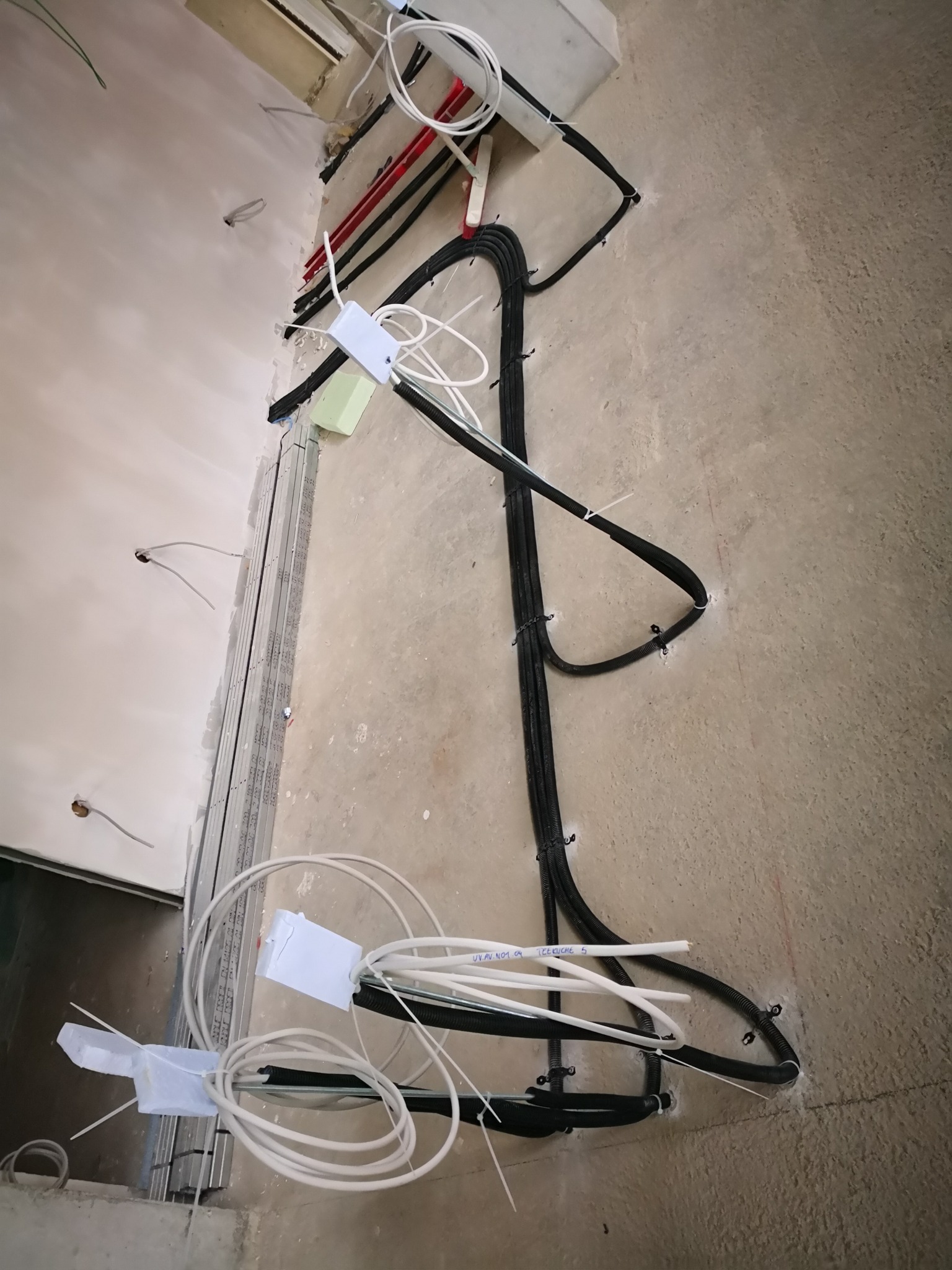 Instalacja elektryczna w trakcie montażu w surowym stanie, widoczne czarne peszle z przewodami, białe kable i puszki instalacyjne zamocowane do ściany, szyny montażowe przy ścianie.