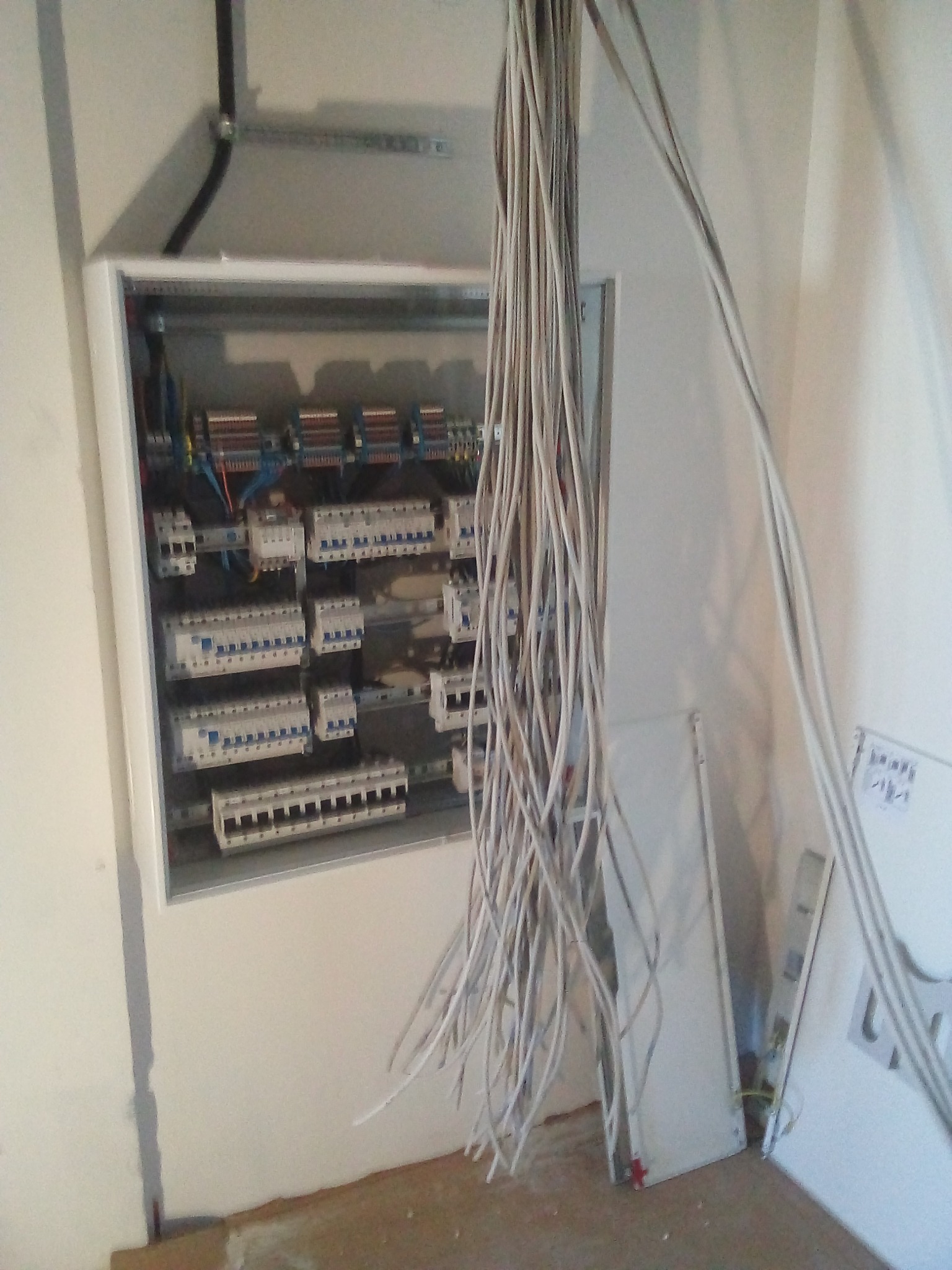 Otwarta skrzynka rozdzielcza z licznymi bezpiecznikami i wystającymi przewodami elektrycznymi, widoczne również fragmenty ścian i podłogi w trakcie remontu.