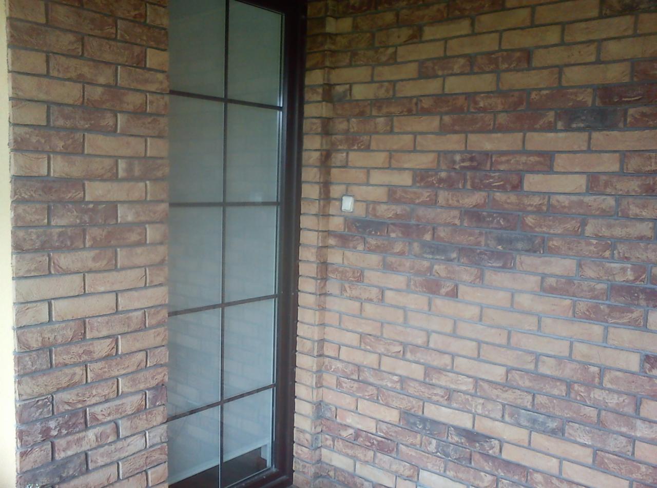 Ściana wykończona cegłą klinkierową w odcieniach brązu i beżu, z widocznym oknem w ciemnej ramie i białym włącznikiem światła.