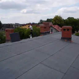 Dach pokryty papą z trzema kominami z czerwonej cegły. Na dachu stoi dekarz z wiadrem, w tle zabudowa jednorodzinna i drzewa.