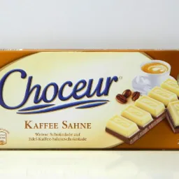 Opakowanie czekolady Choceur Kaffee Sahne z widocznymi kostkami białej czekolady z kawą, obok filiżanka kawy i ziarna kawy.