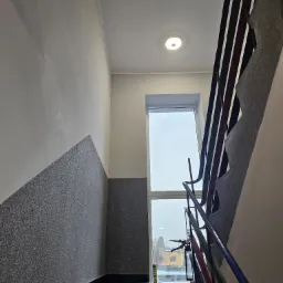 Klatka schodowa w budynku, ściany częściowo wykończone szarą strukturą, częściowo pomalowane na biało, widoczne schody, okno z widokiem na zewnątrz i kolorowe poręcze.