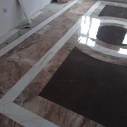 Ukończona podłoga z płytek w odcieniach brązu i bieli, ułożonych we wzór geometryczny z prostokątami i łukami, odbijająca światło, z widocznymi narzędziami i materiałami budowlanymi w tle.