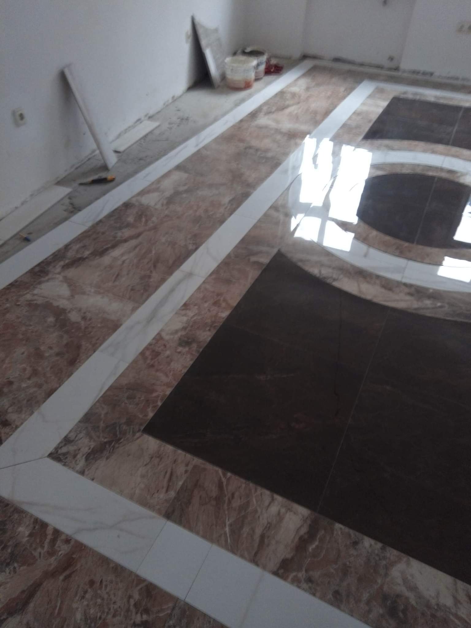 Ukończona podłoga z płytek w odcieniach brązu i bieli, ułożonych we wzór geometryczny z prostokątami i łukami, odbijająca światło, z widocznymi narzędziami i materiałami budowlanymi w tle.