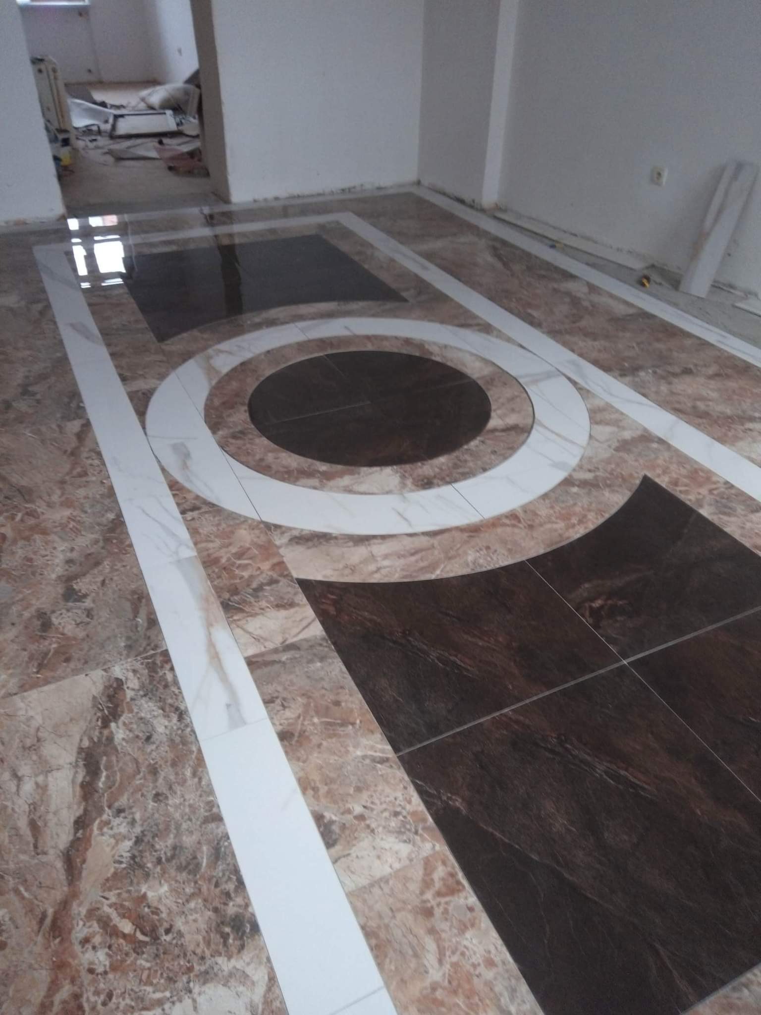 Wzorzysta podłoga z brązowych i beżowych płytek z marmurkowym wzorem, ułożona z białymi płytkami w kształcie obramowania i okręgów, widoczne ślady prac remontowych w tle.